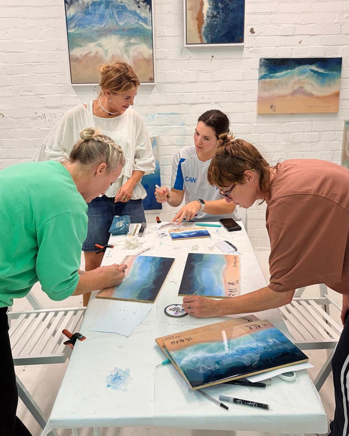 Four people gathered around a white table in an art studio, examining and painting blue and beige landscape scenes. Several framed abstract and landscape paintings are hanging on the white brick wall behind them.