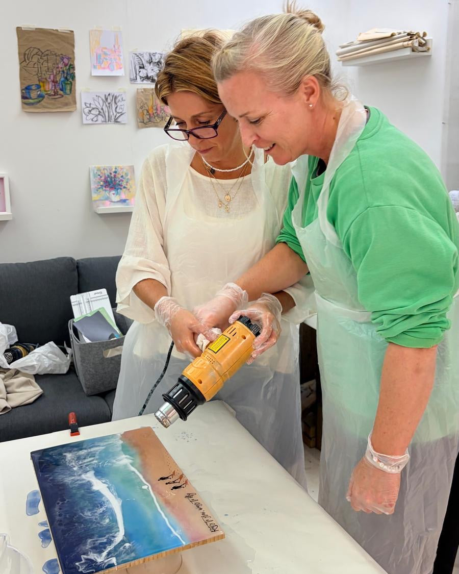 Two women in a studio setting, one wearing a green shirt and apron while the other wears a cream shirt and apron, using a heat gun on a canvas painting of a beach and ocean.
