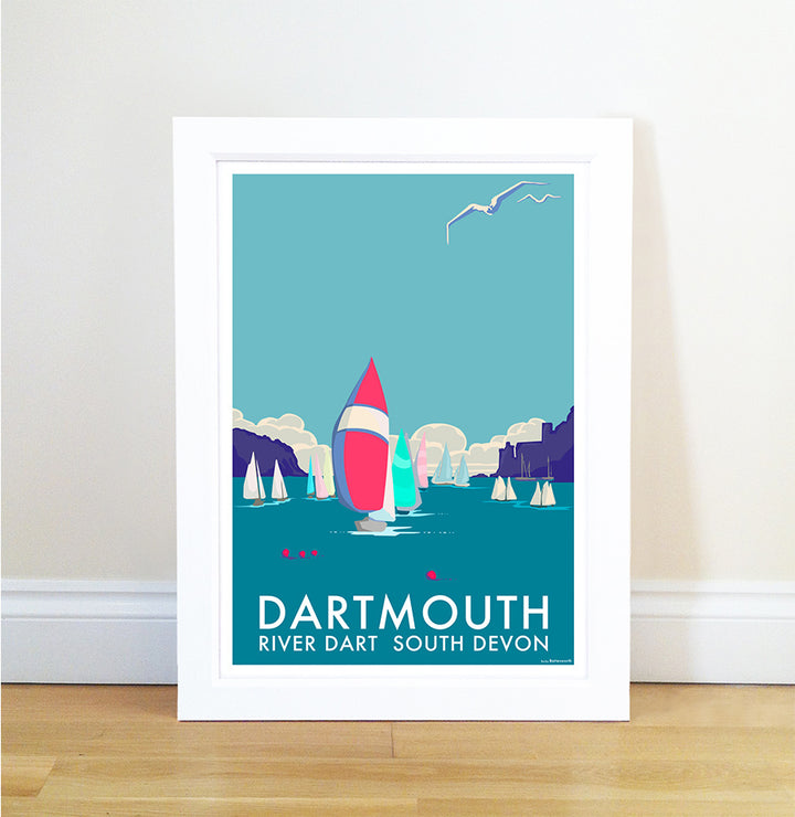 The Dartmouth poster by Becky Bettesworth showcases a vibrant coastal scene with sailboats on blue waters, cliffs, and a clear sky. A seagull glides above the Dartmouth River Dart South Devon text. Its framed and rests on a wooden floor against a white wall.