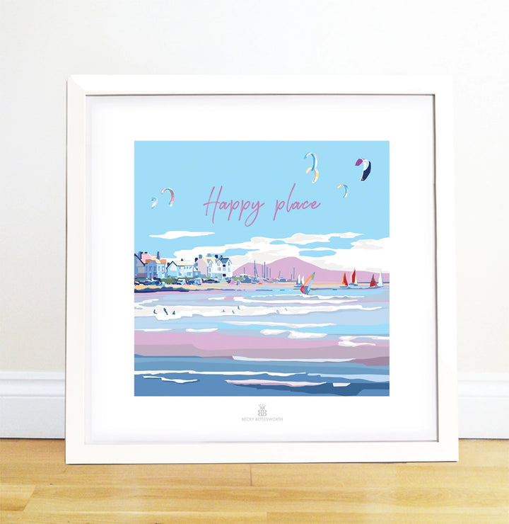 The Happy Place artwork by BeckyBettesworth includes a coastal scene of colorful sailboats, seagulls, and kites under a blue sky. The vintage-style phrase Happy place in pink adorns the top. It stands framed against a white wall on a light wood floor.