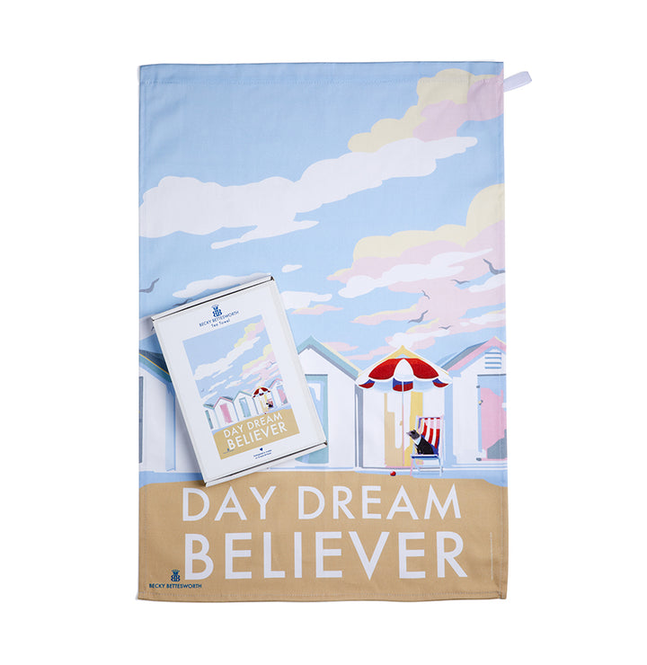 BeckyBettesworths gift-boxed tea towel and notepad feature a charming beach hut scene under a pastel sky, with DAY DREAM BELIEVER text. The tea towel adds artful flair to your kitchen, the design extending into the red-striped umbrella and chair motif on the notepad.