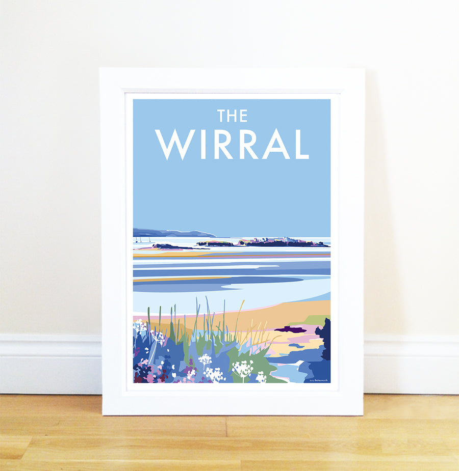 The Wirral framed poster by Becky Bettesworth showcases a vintage seaside print featuring coastal scenery with blue skies, distant hills, water, and shoreline vegetation. The title THE WIRRAL is displayed on top. It rests against a white wall on a wooden floor.