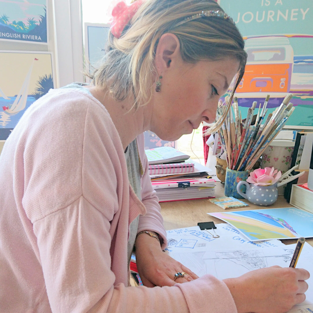 A person in a pink sweater is drawing at a desk with papers and art supplies. Sunlight shines through the window, highlighting colorful posters and a mug of brushes nearby, while Beckys Signature from BeckyBettesworth adds a personal touch to every piece.